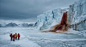 A wide view of explorers looking at the giant red waterfall known as Blood Falls flowing from a white glacier in Antarctica.